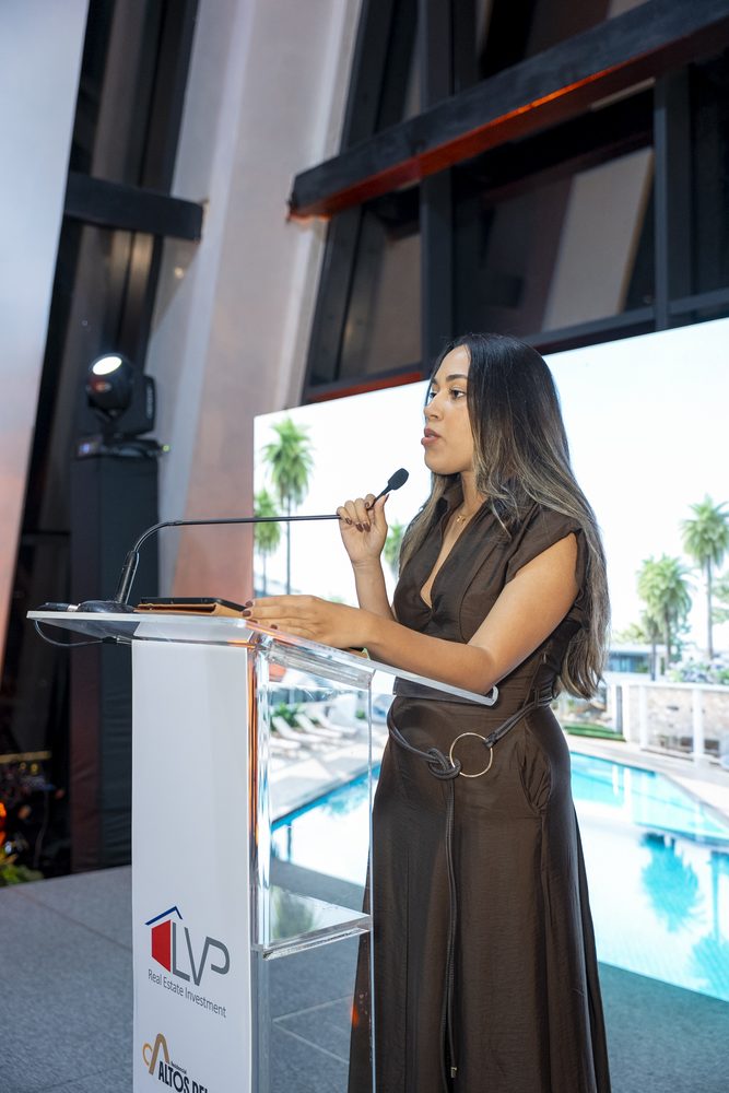 Presentadora hablando en el podio del lanzamiento con la piscina del proyecto al fondo.