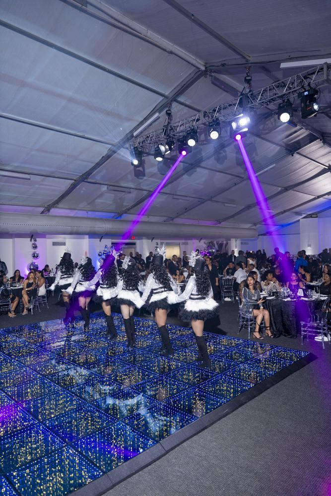 Cuerpo de baile en escena mientras haces de luz violeta cruzan el salón.
