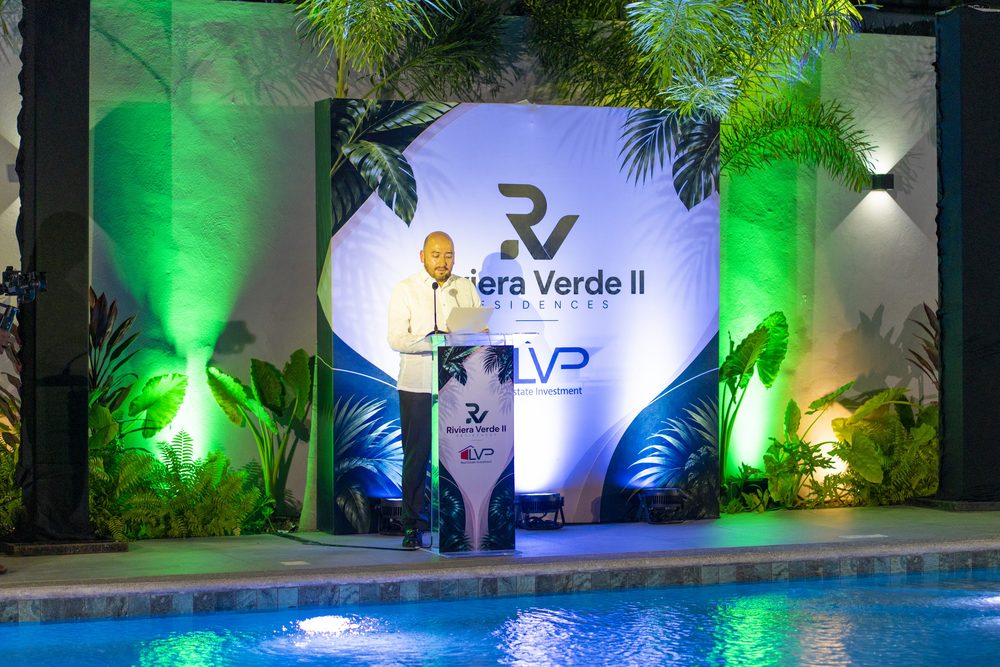 Orador en el podio ante el backdrop de Riviera Verde II, con la piscina iluminada en verde.